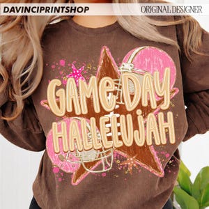 May include: A brown crewneck sweatshirt featuring a graphic print of a brown football, a pink football helmet, and pink stars. The text "GAME DAY HALLELUJAH" is printed in large, light-colored letters across the center of the design.