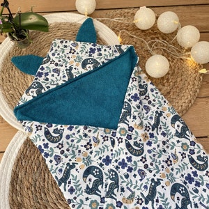 Peut inclure: Une serviette à capuche turquoise avec un motif floral blanc et bleu représentant des renards et des oiseaux. La capuche est en forme de tête de renard.