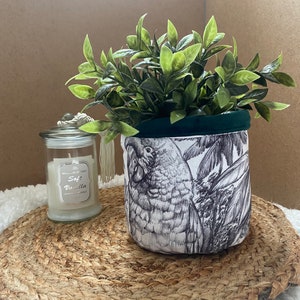 Peut inclure: Un pot en tissu blanc avec un motif de perroquet noir et blanc. Le pot a un dessus en tissu vert et est rempli d'une plante verte feuillue. Le pot est posé sur un tapis brun tissé.