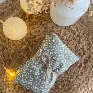May include: Two small, rectangular pillows with a green floral pattern tied together with twine. The pillows rest on a woven, circular mat, with string lights and a white vase in the background.