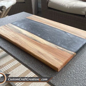 May include: A large wooden cutting board with a gray epoxy resin inlay. The board has a natural wood grain pattern and is finished with a clear coat.