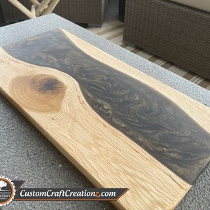 May include: A rectangular wooden cutting board with a dark gray epoxy resin inlay. The resin has a swirling pattern and is shaped like a river flowing through the center of the board.