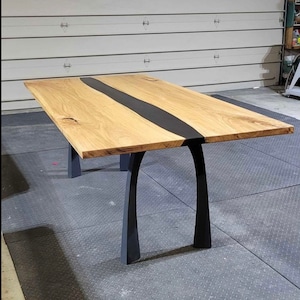 Könnte beinhalten: Ein Esstisch aus Holz mit einem schwarzen Epoxidharzfluss, der durch die Mitte verläuft. Der Tisch hat schwarze Metallbeine mit einem einzigartigen Bogen-Design.