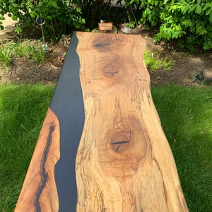May include: A long, wooden table top with a live edge and a black epoxy river running down the center. The table is supported by two black metal legs.