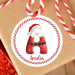 Puede incluir: Una caja de regalo marrón atada con una cuerda roja y blanca. Una pegatina redonda con la letra "A" con temática de Papá Noel y el nombre "Amelia" está adjunta. La pegatina tiene un borde a rayas rojas y blancas.
