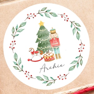 Könnte beinhalten: Ein runder Weihnachtsgeschenkanhänger mit einer Aquarellillustration eines Weihnachtsbaums, eines Nussknackers, eines Schaukelpferds und einer Trommel, umgeben von einem Kranz aus grünen Blättern und roten Beeren. Der Name "Archie" ist geschrieben.