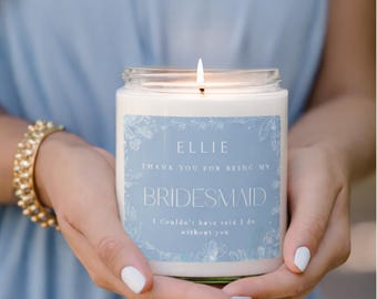 May include: A burning candle in a clear glass jar with a light blue label. The label says "ELLIE THANK YOU FOR BEING MY BRIDESMAID" with extra text. The candle is held in two hands, with a gold bracelet visible.