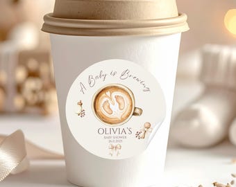 May include: A white takeaway coffee cup with a brown lid. A circular sticker on the cup displays a coffee cup design with baby footprints and the text "A Baby is Brewing" and "Olivia's Baby Shower 26.11.2025".