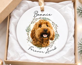 May include: A round white ceramic ornament featuring a portrait of a brown curly-haired dog inside a green pine wreath. The text Bonnie is printed above the dog, and the text Forever Loved is printed below, suspended by a gold ribbon.
