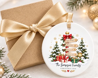 May include: A round white ceramic Christmas ornament featuring a central signpost with four wooden planks labelled Mummy, Daddy, Gracie, and Lucas. The design includes two pine trees, wrapped gifts, and the text The Simpson Family Christmas 2026.