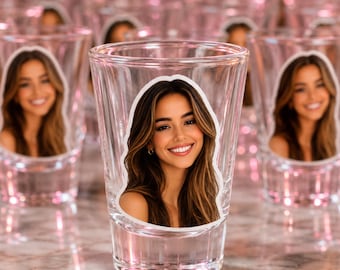 May include: A clear glass shot glass featuring a custom photo decal of a smiling person with long brown hair. The shot glass is part of a larger collection of identical glasses arranged in rows, creating a repetitive visual pattern.
