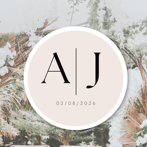 Peut inclure: Un design circulaire avec les initiales "A" et "J" en noir, séparées par une ligne verticale, sur un fond beige clair. La date "02/08/2026" est en dessous des initiales. Le fond présente des compositions florales.