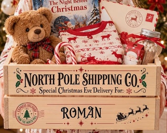 May include: A wooden crate labelled North Pole Shipping Co. with the name Roman printed on the front. Inside are a brown teddy bear with a plaid bow, a copy of The Night Before Christmas, a red and white pyjama set, a candy cane, and a jar of cocoa.