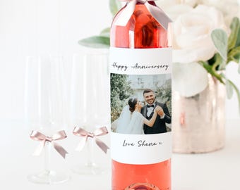 May include: A bottle of rosé wine with a white label featuring a photo and the text "Happy Anniversary" and "Love Shana & ". The bottle is decorated with a pink ribbon. Two champagne flutes with pink bows are in the foreground.
