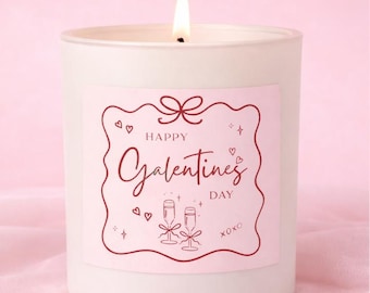 May include: A white candle with a lit wick and a pink label. The label says "Happy Galentine's Day" in cursive, with heart and champagne flute illustrations. The candle sits on a pink surface.