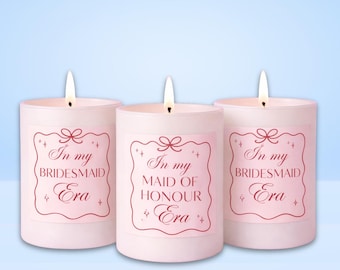 May include: Three pink candles with lit wicks, each bearing a label with text in a decorative font. The labels read "In my BRIDESMAID Era" and "In my MAID OF HONOUR Era". The background is a light blue gradient.