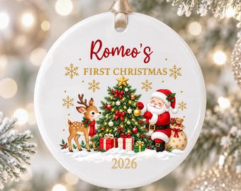 May include: A round white ceramic Christmas ornament hanging from a beige ribbon. The design features a reindeer, a decorated Christmas tree, Santa Claus, and gifts. Text reads: Romeo's FIRST CHRISTMAS 2026, accented with gold snowflakes.