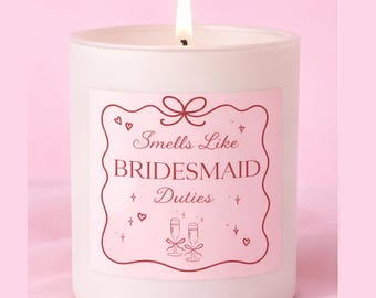 May include: A white candle with a pink label that reads "Smells Like Bridesmaid Duties." The label features a decorative border with hearts, bows, and champagne glasses. The candle is in a white glass jar, and the background is pink.