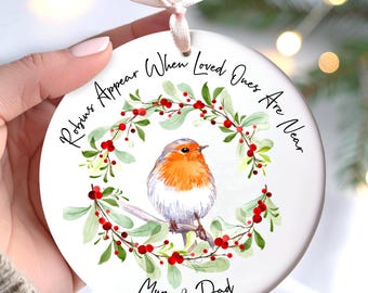 May include: A round white ceramic ornament featuring a watercolour illustration of a robin perched on a branch within a wreath of green leaves and red berries. The text reads "Robins Appear When Loved Ones Are Near" and "Mum & Dad".