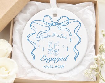 May include: A round white ceramic ornament featuring a blue line-art design of a bow, two champagne flutes, and the text Sophie & Luke Engaged 12.05.2026. The ornament hangs from a white ribbon and is nestled inside a square cardboard gift box.