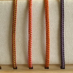May include: Four colorful braided bracelets displayed on a wooden background. The bracelets are in shades of orange, pink, orange, and purple.