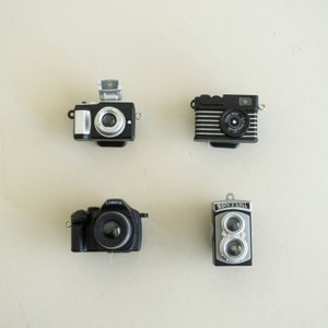 May include: Four miniature cameras in black and silver with different styles. The cameras are on a white surface.