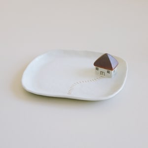 May include: A small white ceramic house with a brown roof sits on a white ceramic dish. The dish has a gold line design that resembles a path leading to the house.