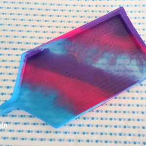 May include: A rectangular silicone mold with a house shape. The mold is filled with a blue, pink, and purple resin mixture. The resin has a cloudy, swirled appearance.