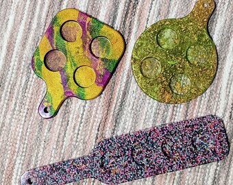 May include: Three different resin serving trays with a glitter finish. The trays are shaped like a paddle, a circle, and a square. The paddle tray is purple and black with a glitter finish. The circle tray is green and gold with a glitter finish. The square tray is green, purple, and gold with a glitter finish.