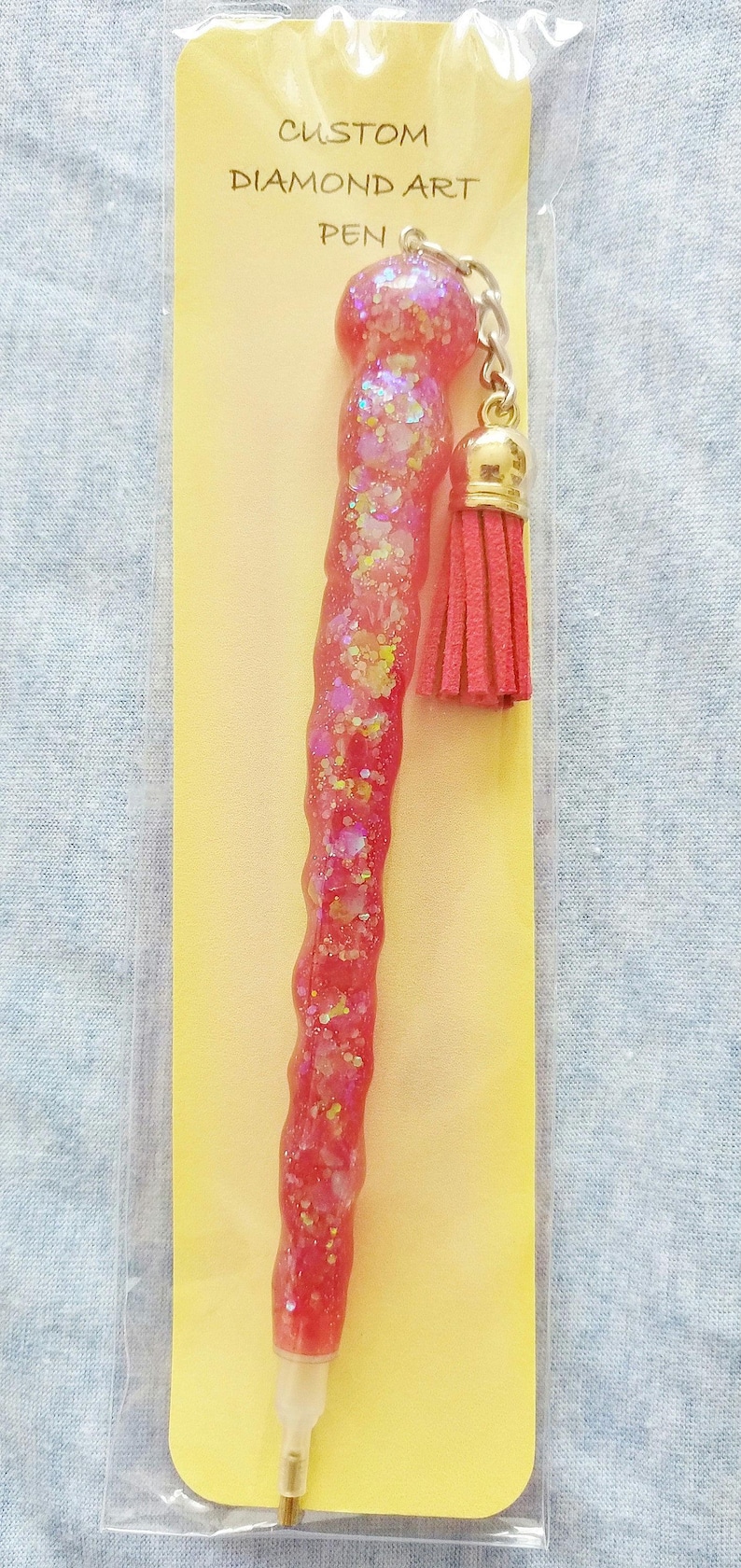 May include: A pink diamond art pen with a gold tassel. The pen is packaged on a yellow card with the text "CUSTOM DIAMOND ART PEN".