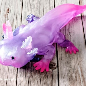 May include: A detailed resin axolotl figurine, predominantly purple with pink accents. The sculpture has intricate features, including gills and tiny feet. The axolotl is displayed on a wooden surface, highlighting its iridescent finish and unique design.
