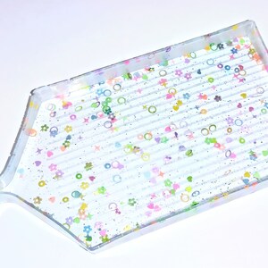 May include: A clear plastic diamond painting tray with a textured surface and a handle. The tray is decorated with colorful glitter and small shapes, including hearts, stars, and circles.