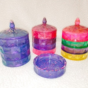 May include: Three tiered decorative containers with lids. The containers are made of clear resin and have a sparkly finish. The top tier is pink, the middle tier is green, and the bottom tier is yellow. The container on the left is purple and has a matching tray.