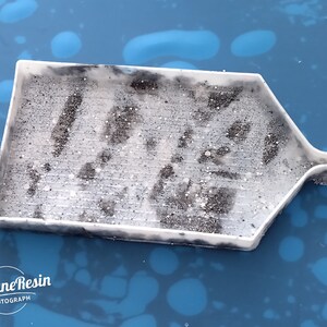 May include: A white plastic diamond painting tray with a textured surface and a small spout on the side. The tray is filled with black diamond painting drills.