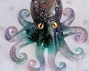 May include: A handcrafted octopus figurine with a glittery finish. The octopus features a dark purple head, transitioning to teal and lavender tentacles. The eyes are a bright yellow-orange. The figurine is set against a white background.