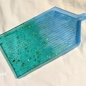 May include: A blue and green resin tray with a textured surface and glitter embedded in the resin. The tray has a handle on the top right corner.