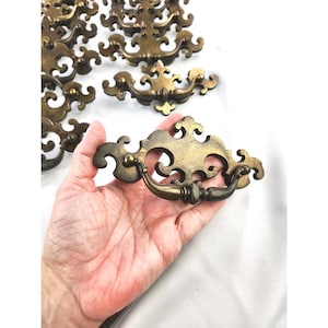 May include: A collection of brass-toned metal drawer pulls featuring an ornate, symmetrical backplate with intricate scrollwork and a curved, swinging handle. The hardware has a weathered, aged finish with visible dark patina in the crevices.
