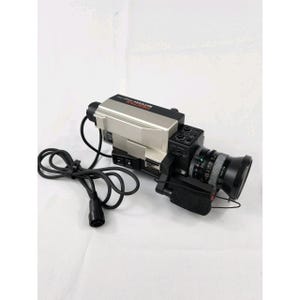 May include: A silver and black Pentax Saticon video camera featuring a lens, a side-mounted handle, and a long black power cable with a multi-pin connector. The top of the camera body displays the brand name Pentax and the word Saticon.