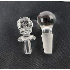 May include: Two clear glass bottle stoppers. One stopper has a crown design on top, and the other has a round top. Both stoppers have a tapered base for insertion into a bottle.