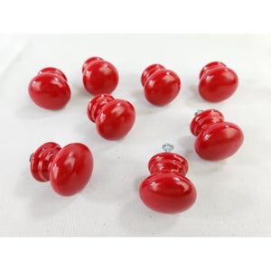 May include: A collection of eight round, glossy red cabinet knobs. Each knob features a smooth, spherical top surface and a stepped base design. A small silver-colored metal screw is visible protruding from the center of the base on each knob.