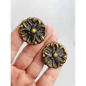 May include: Two circular metal buttons featuring a raised, ornate floral relief pattern with a dark, antiqued brass finish. Each button has a central circular dome surrounded by symmetrical, curved leaf-like motifs and a scalloped outer edge.