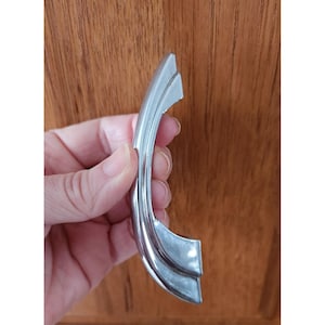 May include: A curved, chrome-colored cabinet handle with a sleek, modern design. The handle is held in a hand, showcasing its polished finish and curved shape. The handle is approximately 6 inches long.