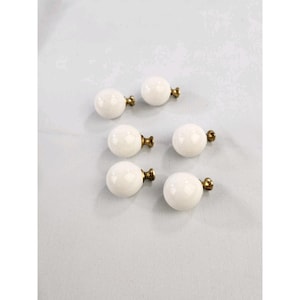 May include: A set of five spherical white knobs, each featuring a smooth, glossy finish and a metallic brass-toned base. These round cabinet pulls are arranged in a cluster, showcasing their uniform shape and the contrast between the white surface.