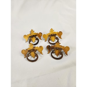 May include: A set of four gold-toned metal drawer pulls featuring a decorative backplate with a leaf-like shape and a dark, circular drop handle. Each piece displays a weathered finish with visible signs of oxidation and patina on the metal surface.