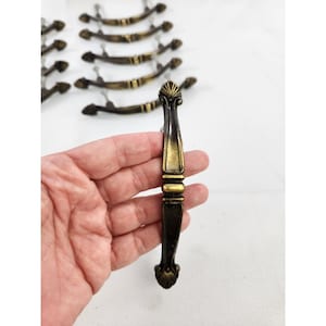 May include: A collection of antique-style metal drawer pulls featuring a dark bronze finish with gold-toned highlights. Each handle has a central decorative band and shell-like motifs at both ends, with a curved shape for easy gripping.