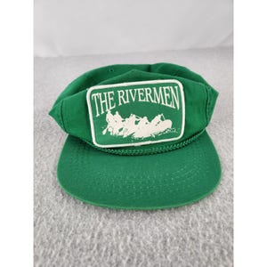 May include: A green baseball cap featuring a rectangular patch on the front. The patch displays the white text THE RIVERMEN above a graphic illustration of people in a raft. A green braided cord runs across the base of the crown above the brim.