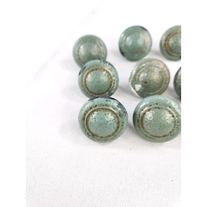 May include: A collection of round, sage green buttons featuring a textured, speckled surface and a raised circular rim detail. Each button has a slightly weathered appearance with subtle metallic-toned accents along the edges and center.