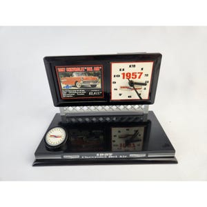 May include: A black desk clock featuring a 1957 Chevrolet Bel Air theme. The clock has a rectangular face with a picture of the car and the year 1957. A smaller clock is on the base. The clock has a silver bridge design.