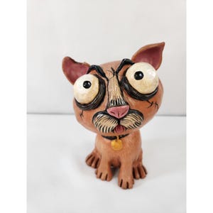 May include: A brown cat figurine with oversized, bulging white eyes, black pupils, and thick black outlines around the eyes. The cat has a pink nose, a small tongue sticking out, a black collar with a gold tag, and visible stitch-like markings.