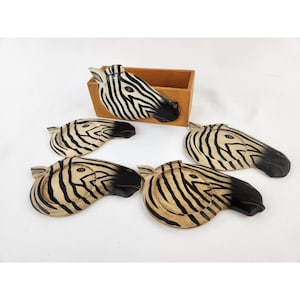 May include: A set of four wooden coasters shaped like zebra heads with black and cream stripes, accompanied by a rectangular wooden storage box. Each coaster features a carved zebra face with a black muzzle and detailed eye markings.